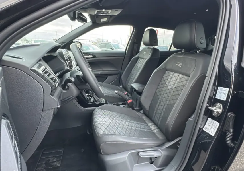 Vue intérieure côté conducteur d'un Volkswagen T-Cross noir intense 2025, sièges tissu R-Line avec surpiqûres blanches.