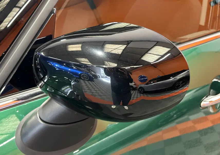 Rétroviseur noir brillant côté gauche d'une MINI Countryman vert métallisé, vue rapprochée sur fond intérieur showroom.