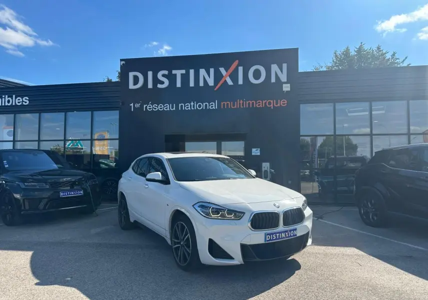 BMW X2 blanc alpin vue 3/4 avant droit avec jantes M 19 pouces devant un showroom Distinxion