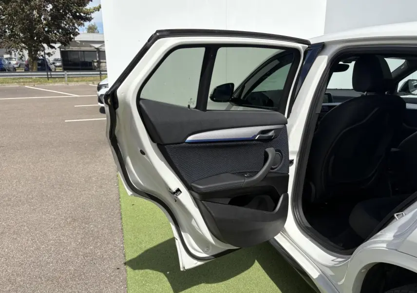 Portière arrière gauche ouverte d'une BMW X2 blanche Alpine, montrant l'intérieur noir avec inserts bleus et poignée intégrée.