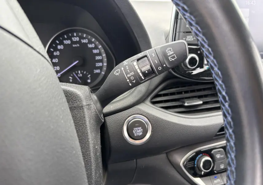 Gros plan intérieur sur le volant avec surpiqûres bleues, bouton start/stop et commandes d'essuie-glace Hyundai i30 SW blanche.