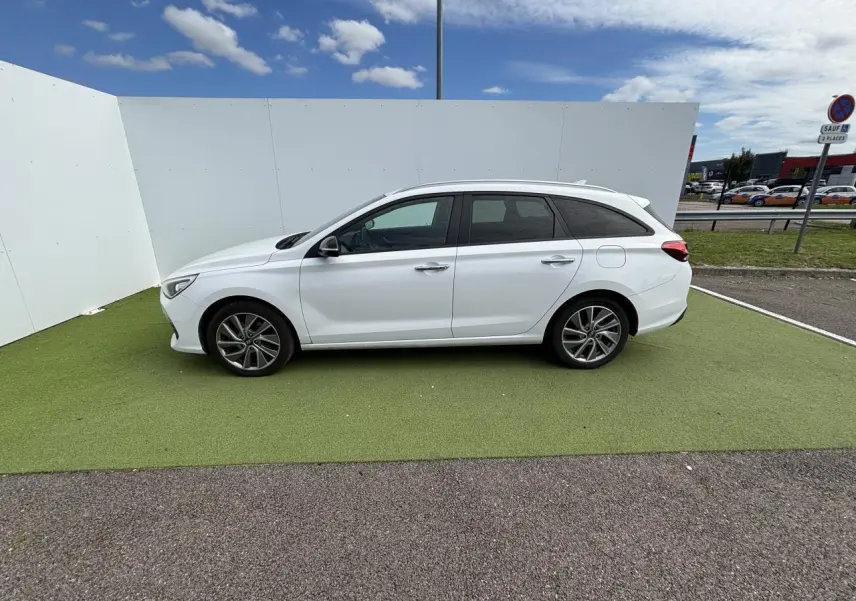 Vue latérale côté gauche d'une Hyundai i30 SW blanche garée sur sol vert avec jantes alliage et vitres surteintées.