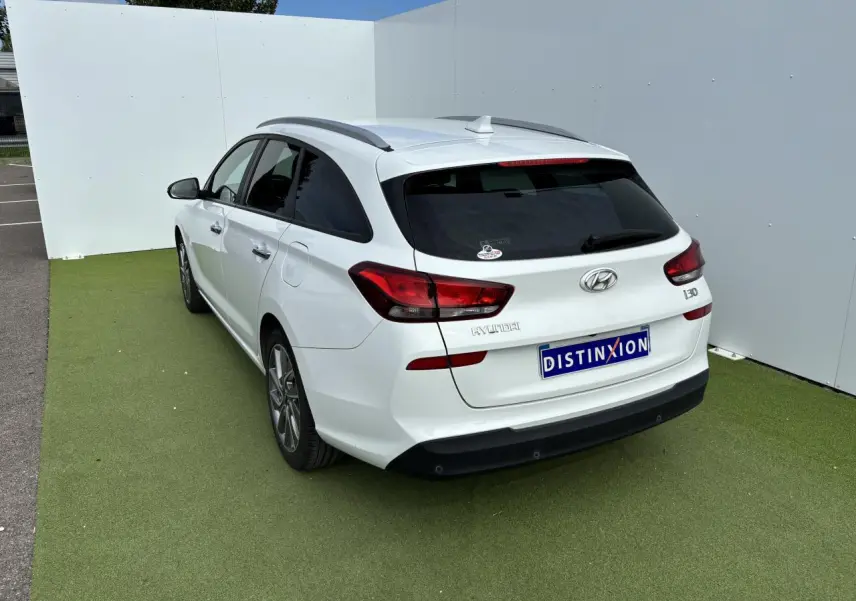 Vue 3/4 arrière droite d'une Hyundai i30 break blanche avec vitres arrière surteintées et barres de toit noires