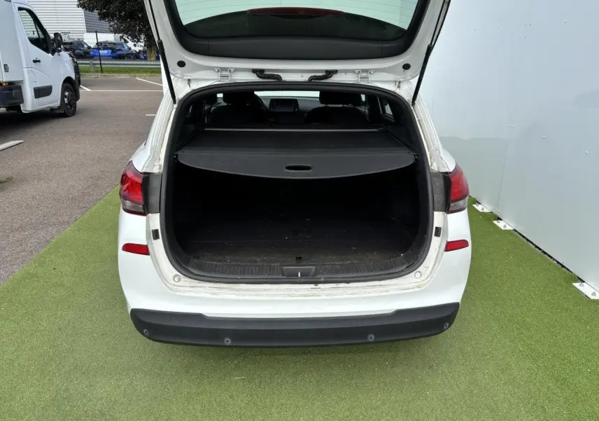 Coffre ouvert vu de l'arrière d'une Hyundai i30 SW blanche, montrant le cache-bagages et l'espace de chargement vide.