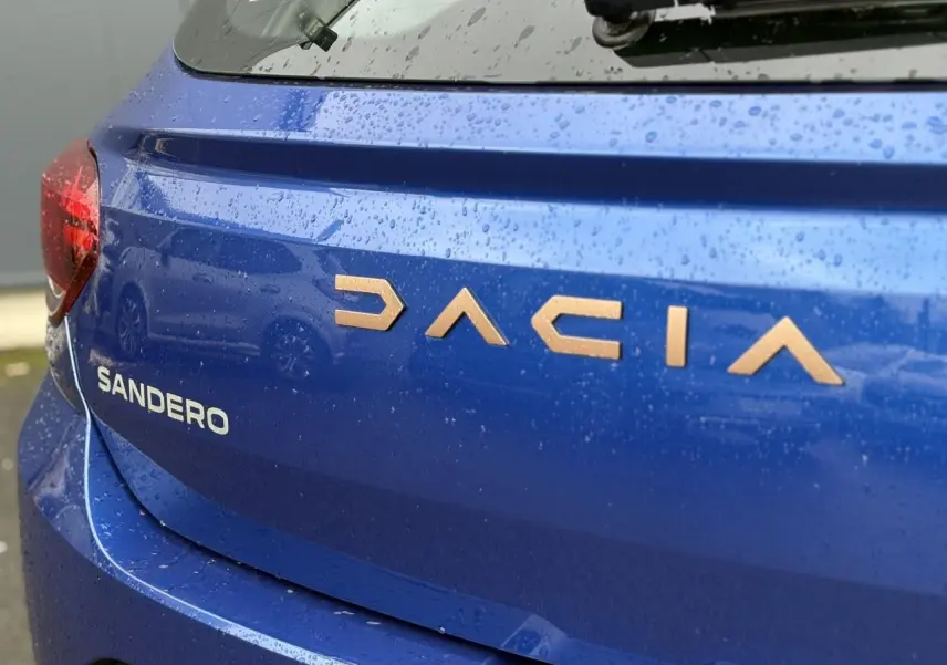 Gros plan arrière droit sur le coffre bleu Iron d'une Dacia Sandero avec lettrage cuivré sous pluie fine.