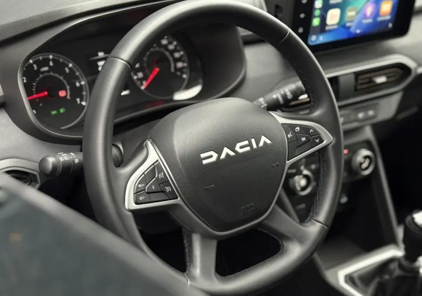 Gros plan sur le volant noir Dacia avec commandes intégrées, tableau de bord et écran tactile du poste de conduite.