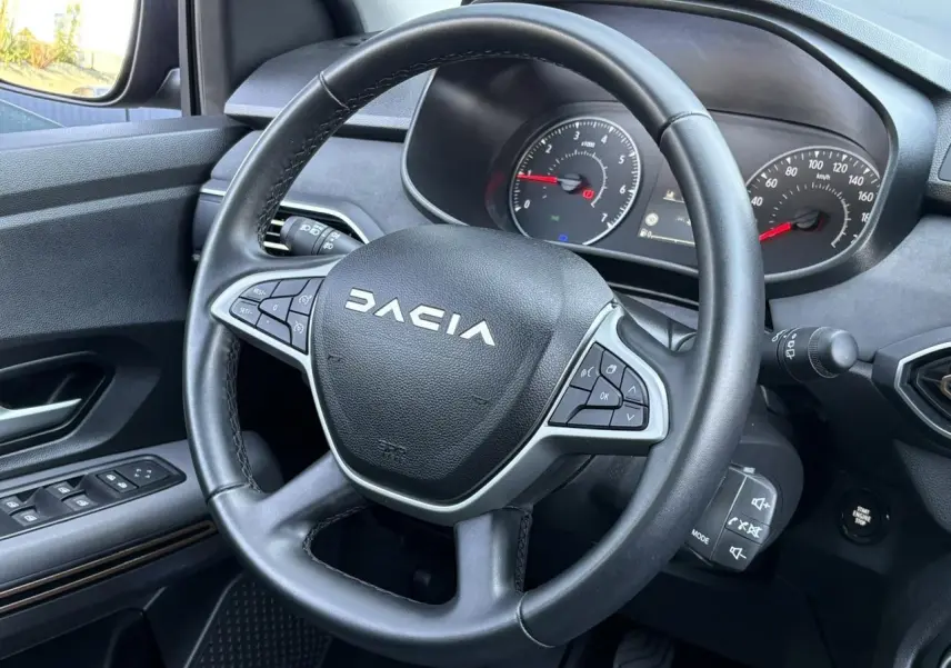Vue intérieure centrée sur le volant noir Dacia avec commandes intégrées et tableau de bord analogique.