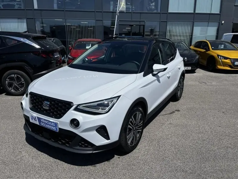 SEAT Arona blanc vue 3/4 avant droit, avec toit noir et jantes alliage, stationné devant un bâtiment vitré.