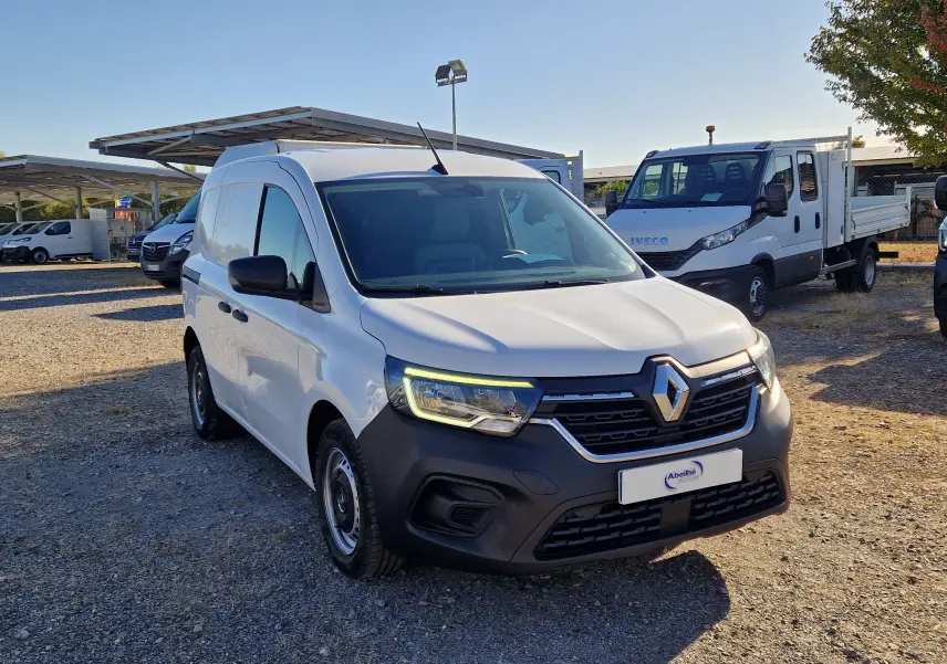 Renault Kangoo blanc en 3/4 avant droit, avec calandre noire et phares allumés sur parking extérieur.
