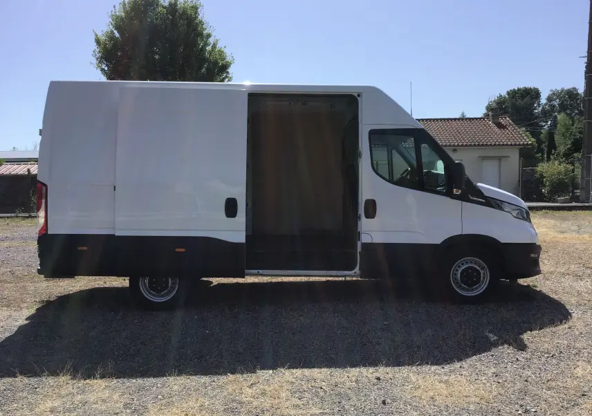 IVECO Daily Fourgon blanc vu de profil côté gauche, porte latérale coulissante ouverte sur un terrain extérieur.