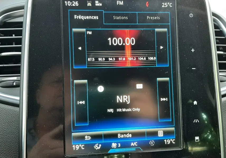Écran central tactile du Renault Scénic IV 2018 affichant la radio NRJ à 100.00 FM avec commandes climatisation en dessous.