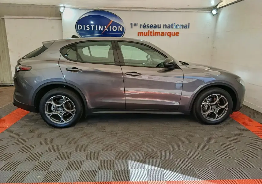 Profil côté gauche de l'Alfa Romeo Stelvio gris foncé 2024 dans un showroom avec jantes spécifiques et logo Distinxion en arrière-plan.