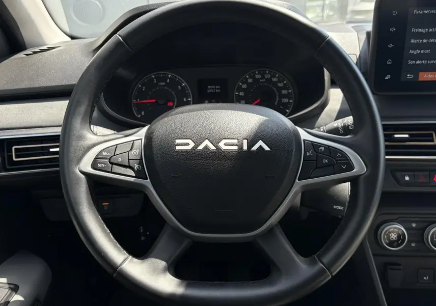 Vue frontale du volant noir DACIA avec commandes intégrées et tableau de bord numérique en arrière-plan.