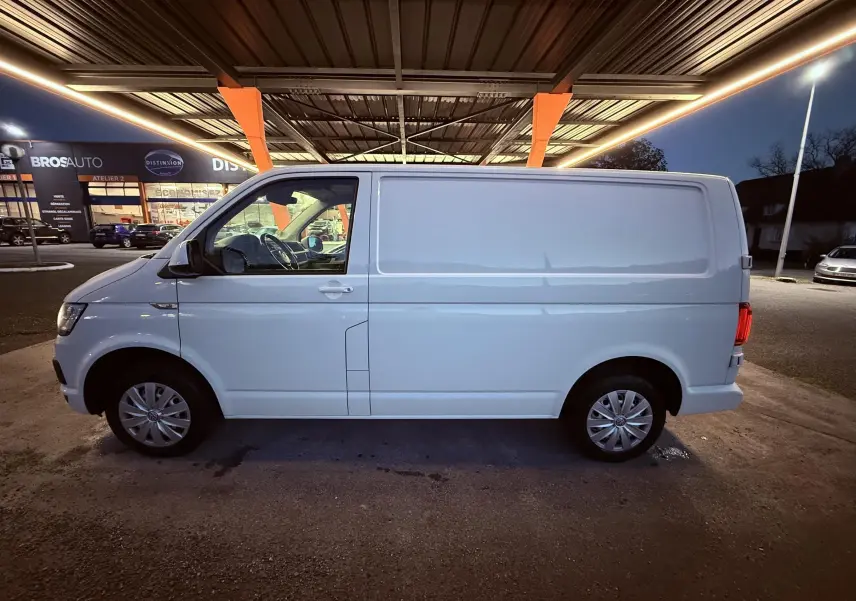 Vue de profil côté gauche d’un Volkswagen Transporter blanc L1H1 2018, stationné sous un auvent éclairé.
