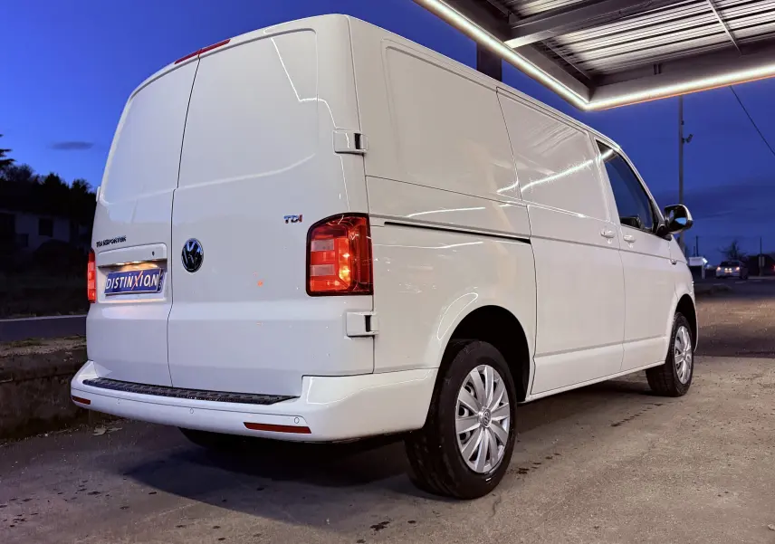 Vue 3/4 arrière droite d’un Volkswagen Transporter L1H1 blanc de 2018, avec feux arrière allumés et jantes argentées.