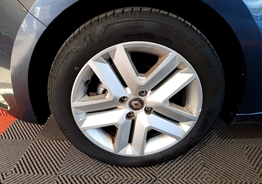 Gros plan sur la roue avant gauche d'une Renault Clio gris foncé, jante alliage au design moderne à cinq branches.