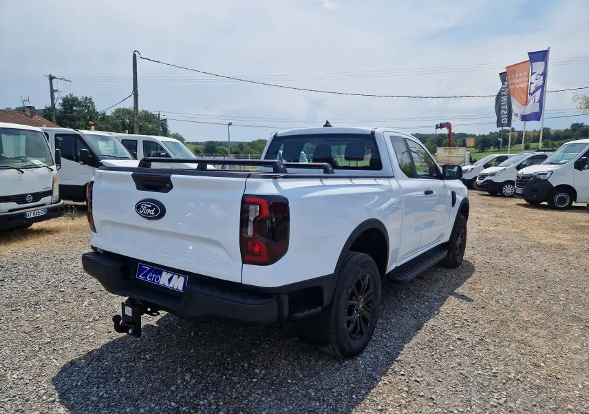 Vue 3/4 arrière droite du Ford Ranger Super Cabine blanc avec plateau, barres de toit et attelage visible sur un terrain gravillonné.