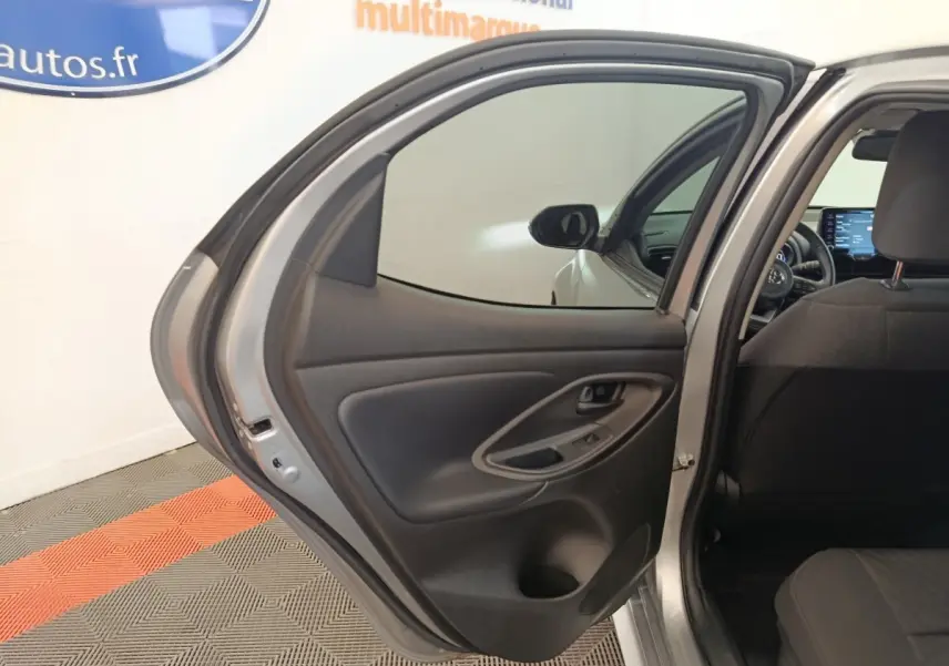 Vue intérieure de la porte arrière gauche ouverte d'une Toyota Yaris hybride gris clair, avec siège arrière et tableau de bord partiellement visibles.