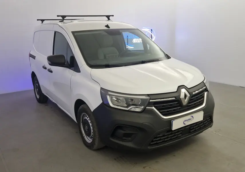 Renault Kangoo blanc en 3/4 avant droit, utilitaire avec calandre noire et barres de toit visibles.
