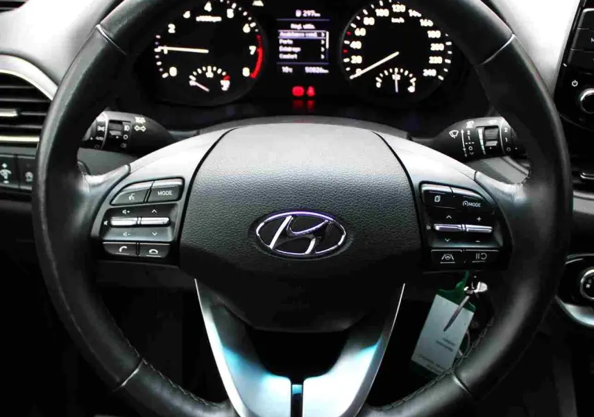 Vue rapprochée du volant noir de la Hyundai i30 SW 2022 avec commandes intégrées et tableau de bord éclairé en arrière-plan.