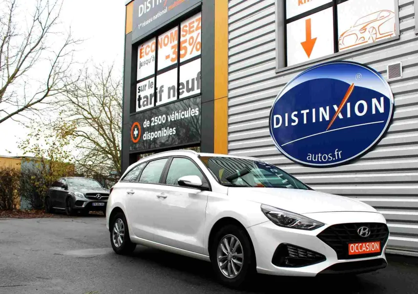 Hyundai i30 SW blanc polaire vue 3/4 avant droit, stationnée devant un bâtiment avec panneau Distinxion.