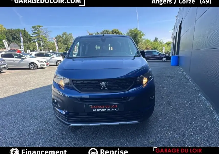 Vue avant d'un Peugeot Rifter bleu 2024 avec calandre noire et feu avant droit allumé sur parking extérieur.