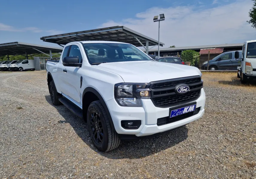 Ford Ranger Super Cabine blanc vu en 3/4 avant droit, avec calandre noire et roues noires sur terrain gravillonné.