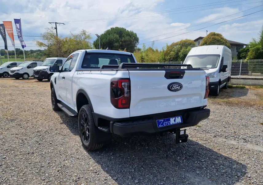 Ford Ranger Super Cabine blanc vu 3/4 arrière droit, avec rideau rigide coulissant et attelage visible sur terrain gravel.