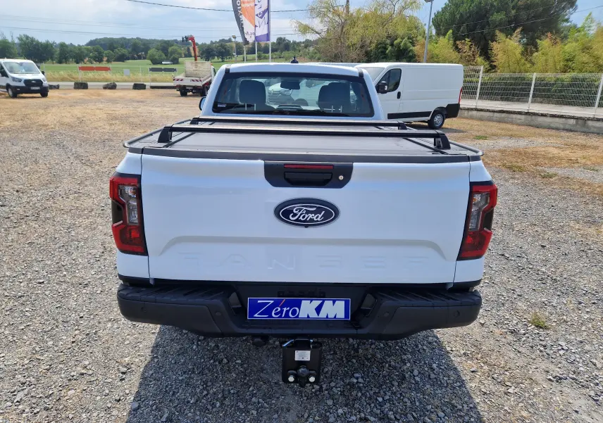 Vue arrière d'un Ford Ranger Super Cabine blanc avec plateau couvert et barres de chargement sur un sol gravillonné.