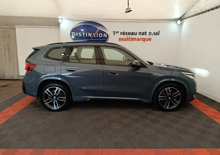 Profil droit d'un BMW X1 xDrive25e M Sport gris foncé, avec jantes alliage et toit spoiler visible en intérieur showroom.