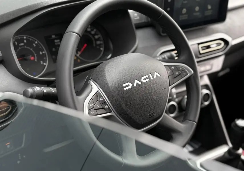 Vue rapprochée du volant DACIA noir avec commandes intégrées et tableau de bord du Jogger 2025 en arrière-plan.