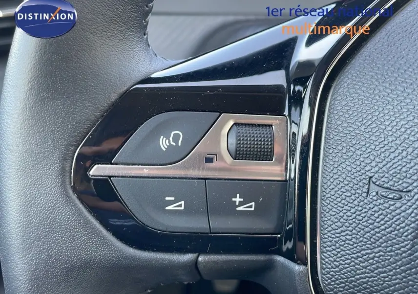 Gros plan sur les commandes de volume au volant noir de la Peugeot 208 blanc banquise, version 2024 essence manuelle.