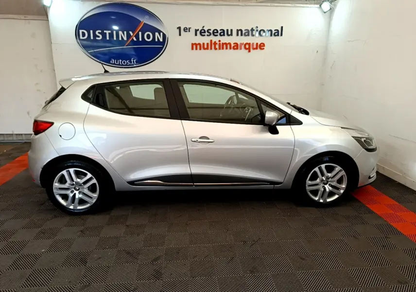 Profil côté droit d'une Renault Clio Business TCe 90 gris clair, en intérieur sur sol damier avec logo Distinxion en arrière-plan.