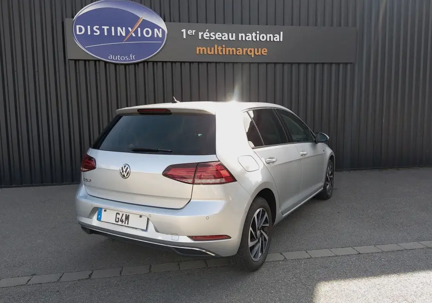 Volkswagen Golf gris reflet d'argent vue 3/4 arrière droit, stationnée devant un mur noir avec enseigne Distinxion.
