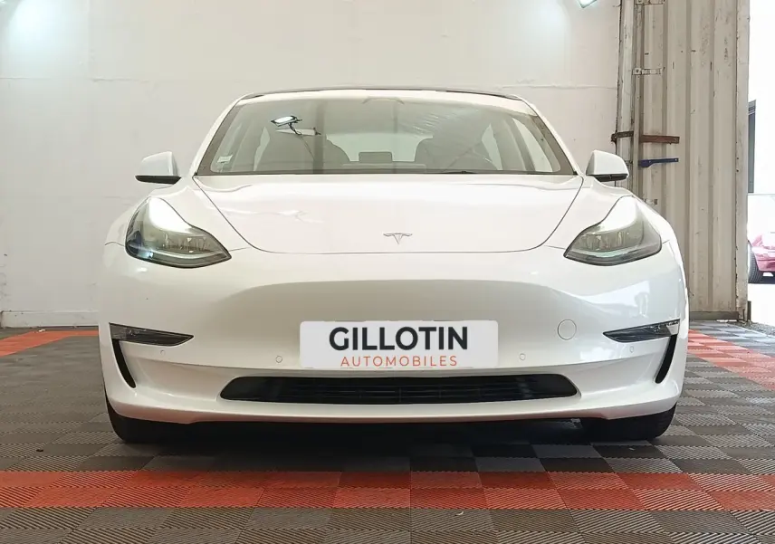 Vue frontale d'une Tesla Model 3 Grande Autonomie blanche, avec phares LED allumés et plaque d'immatriculation GILLOTIN.