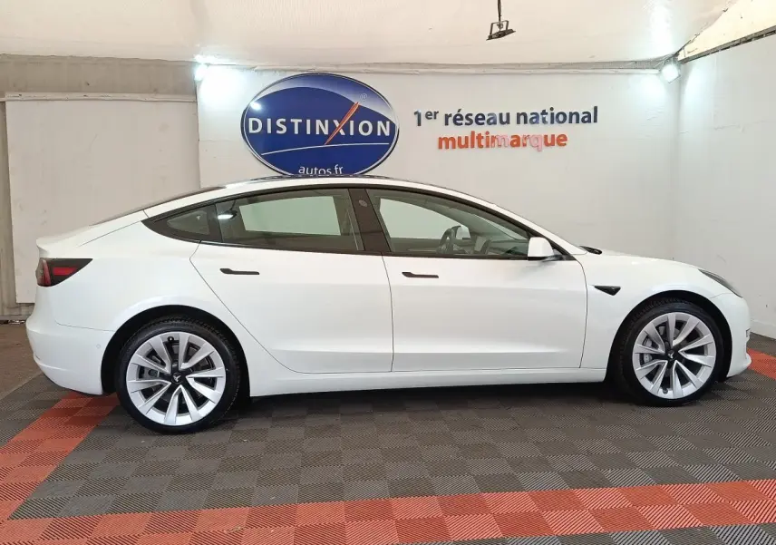 Tesla Model 3 Grande Autonomie blanche vue de profil côté gauche dans un showroom avec sol à damiers rouge et gris.