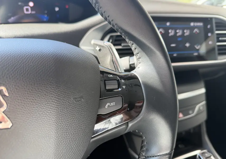 Gros plan sur le volant noir de la Peugeot 308 SW 2021 avec commandes intégrées et tableau de bord numérique flou en arrière-plan.