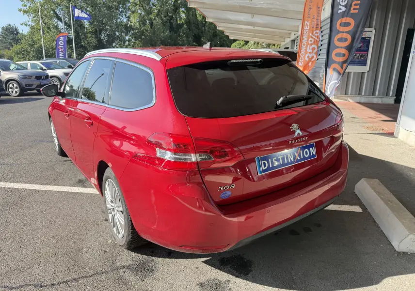 Vue 3/4 arrière droite d'une Peugeot 308 SW rouge rubis stationnée devant un bâtiment avec vitres teintées.