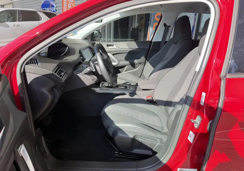 Vue intérieure côté conducteur du Peugeot 308 SW rouge rubis, mettant en valeur les sièges gris et le tableau de bord moderne.