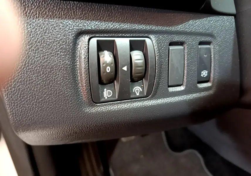 Gros plan sur les commandes de réglage des phares et tableau de bord noir dans une Renault Clio Business blanche de 2019.