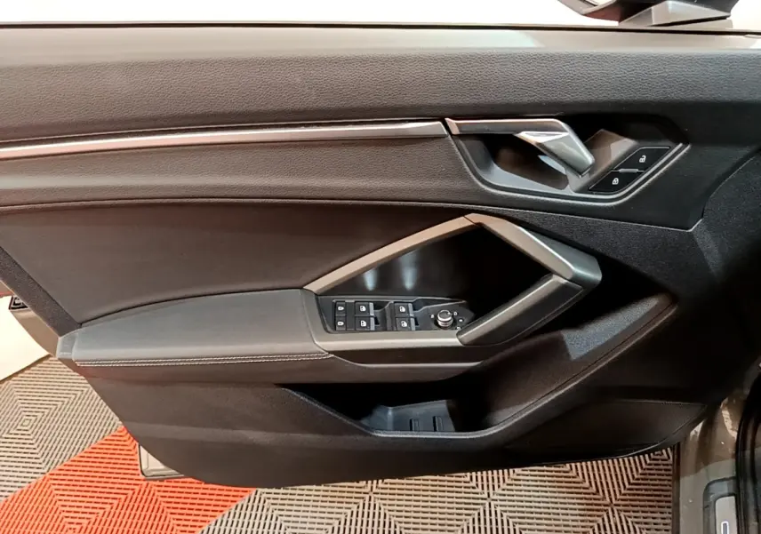 Vue intérieure côté gauche de la porte avant d’un Audi Q3 gris foncé avec commandes de vitres et poignée argentée.