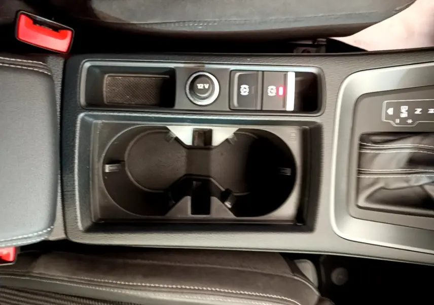 Vue plongeante sur la console centrale noire de l’Audi Q3 35 TFSI 150 MHEV S Line avec porte-gobelets et commandes frein à main électrique.