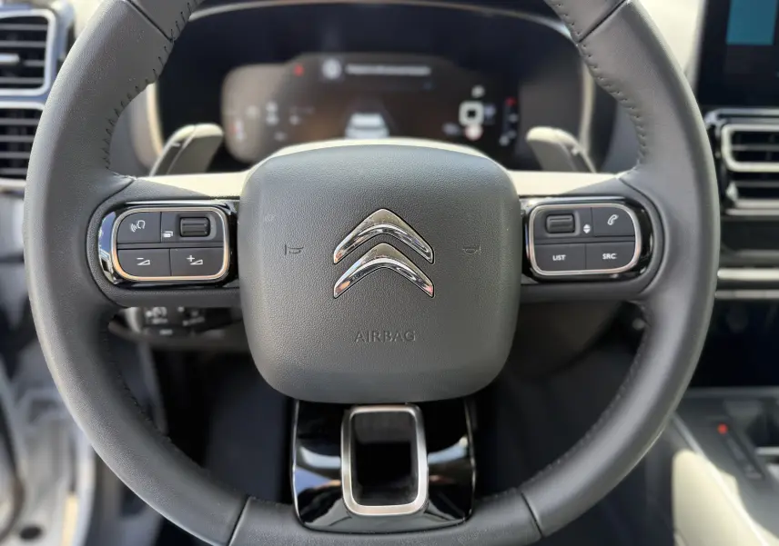 Vue rapprochée du volant noir du Citroën C5 Aircross Hybrid 145 e-DSC6 Max avec commandes intégrées et tableau de bord numérique flou.