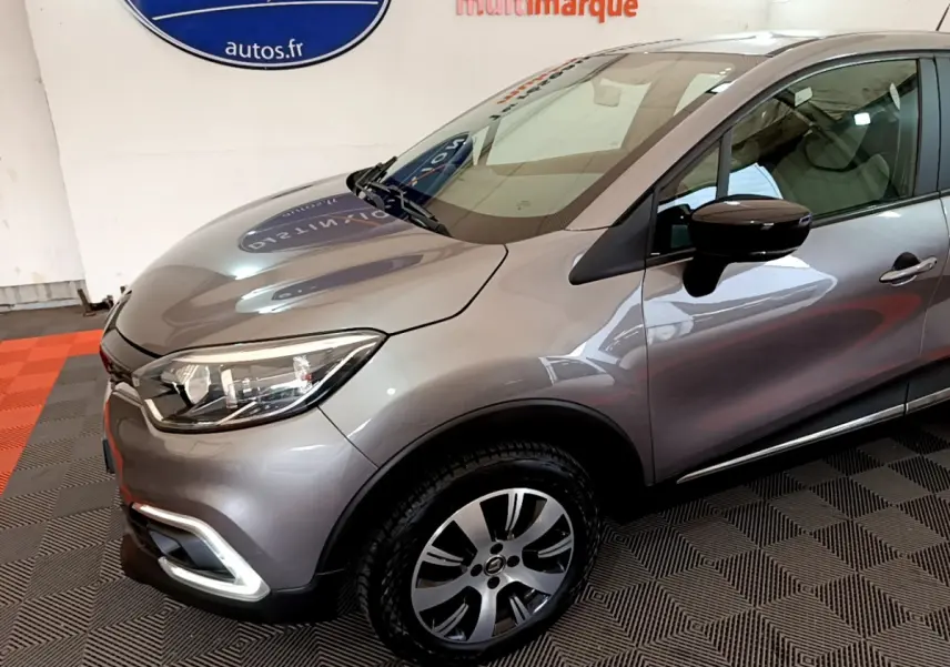 Vue 3/4 avant droit d'un Renault Captur gris foncé avec phares LED et jantes alliage visibles en intérieur showroom.