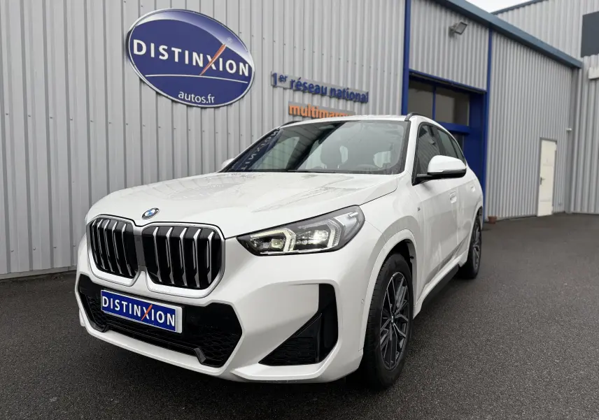 BMW X1 18D blanc minéral en vue 3/4 avant droit, avec calandre imposante et phares LED allumés.