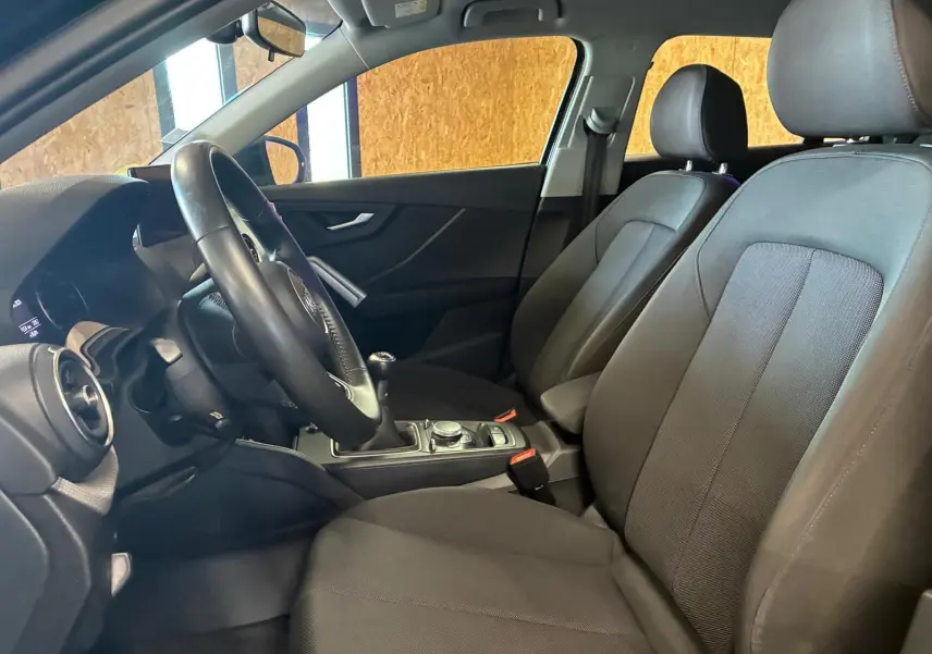 Vue intérieure côté conducteur de l'Audi Q2 noir, montrant les sièges en tissu noir et la console centrale avec boîte manuelle.