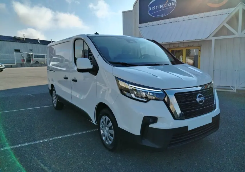 Nissan Primastar blanc en 3/4 avant droit, utilitaire avec calandre noire et détails chromés sous un ciel bleu.