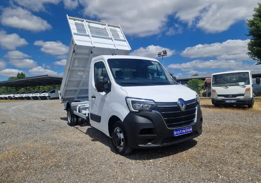 Renault Master Benne blanc en 3/4 avant droit, benne relevée et roues jumelées visibles sur terrain gravillonné.