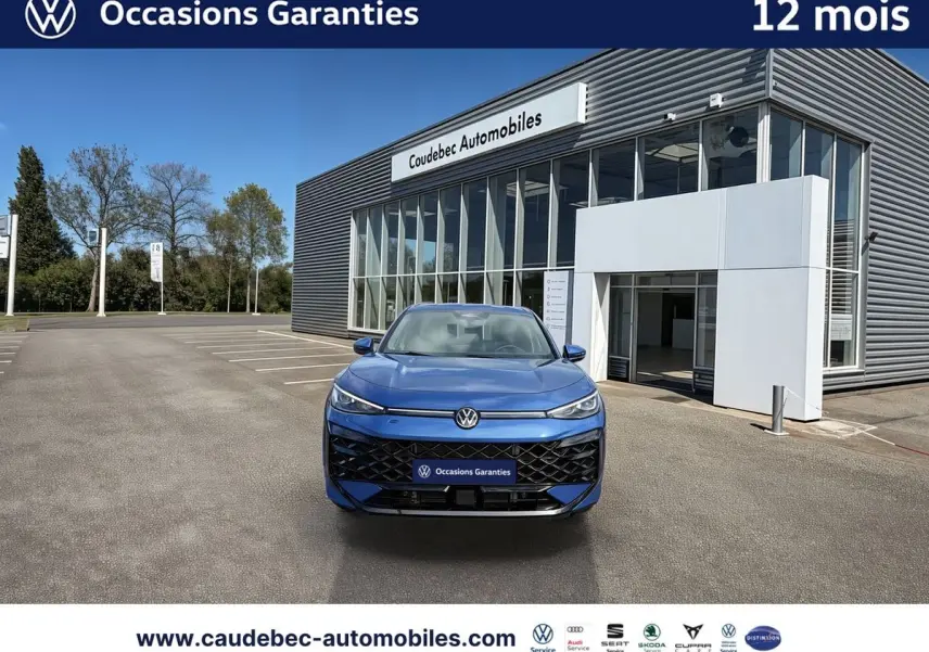 Volkswagen T-Roc bleu métallisé vu de face, avec calandre noire et logo lumineux devant un bâtiment moderne.