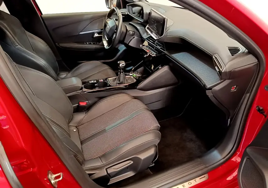 Intérieur côté conducteur d'une Peugeot 208 rouge, montrant sièges noirs, volant cuir et écran tactile central.